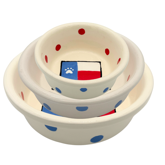 Set of white handmade ceramic dog bowls with Texas flag design and white paw print inside, red polka dots around the interior and blue polka dots around the outside in multiple sizes.